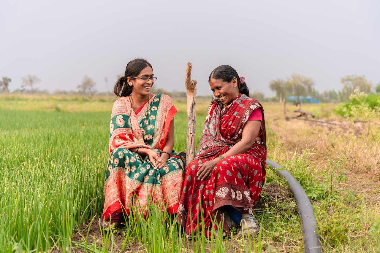 Two smiling Indian women in colorful sarees sitting in a lush green farm, depicting rural life and happiness.